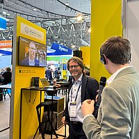 Zwei Männer stehen an einer gelben Stele auf einer Messe und unterhalten sich. Im Hintergrund läuft auf ein Video auf einem Monitor. 