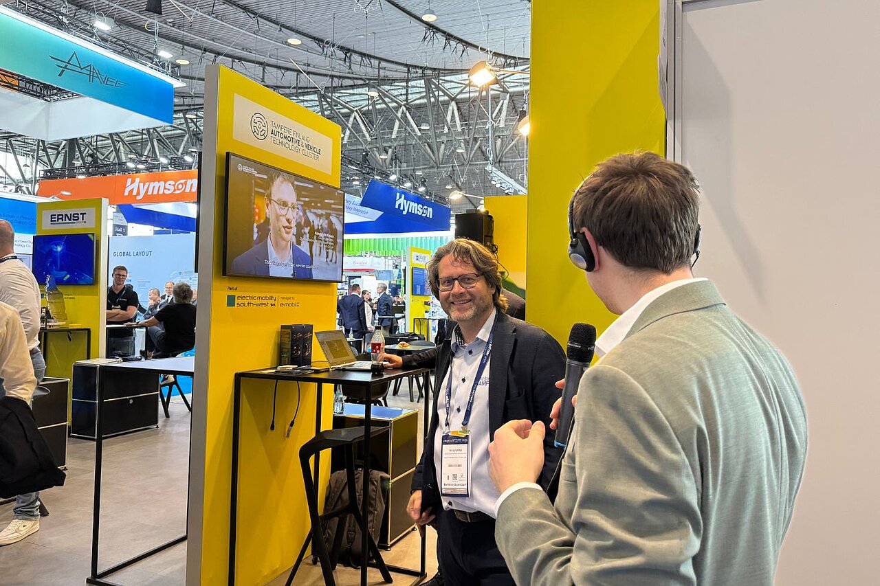 Zwei Männer stehen an einer gelben Stele auf einer Messe und unterhalten sich. Im Hintergrund läuft auf ein Video auf einem Monitor. 