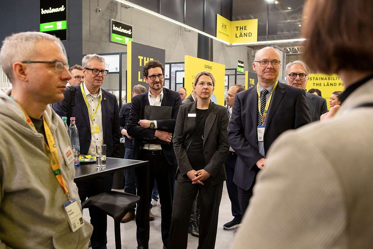 Mehrere Menschen stehen um einen Tisch herum und hören einem Sprecher zu, der nicht zu sehen ist auf dem Bild.