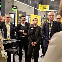 Mehrere Menschen stehen um einen Tisch herum und hören einem Sprecher zu, der nicht zu sehen ist auf dem Bild.