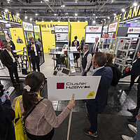 Mehrere Personen stehen um ein Exponat auf der Messe herum, ein Mann erklärt was zu sehen ist.