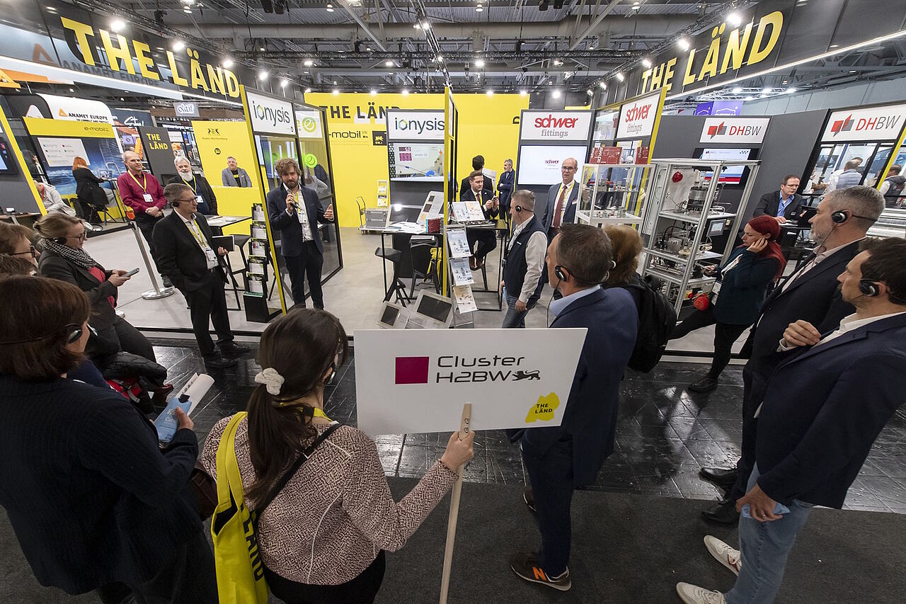 Mehrere Personen stehen um ein Exponat auf der Messe herum, ein Mann erklärt was zu sehen ist.