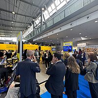 Eine Gruppe Personen informiert sich auf einem Messestand und hört über Kopfhörer einer Führung zu.
