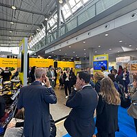Eine Gruppe Personen informiert sich auf einem Messestand und hört über Kopfhörer einer Führung zu.