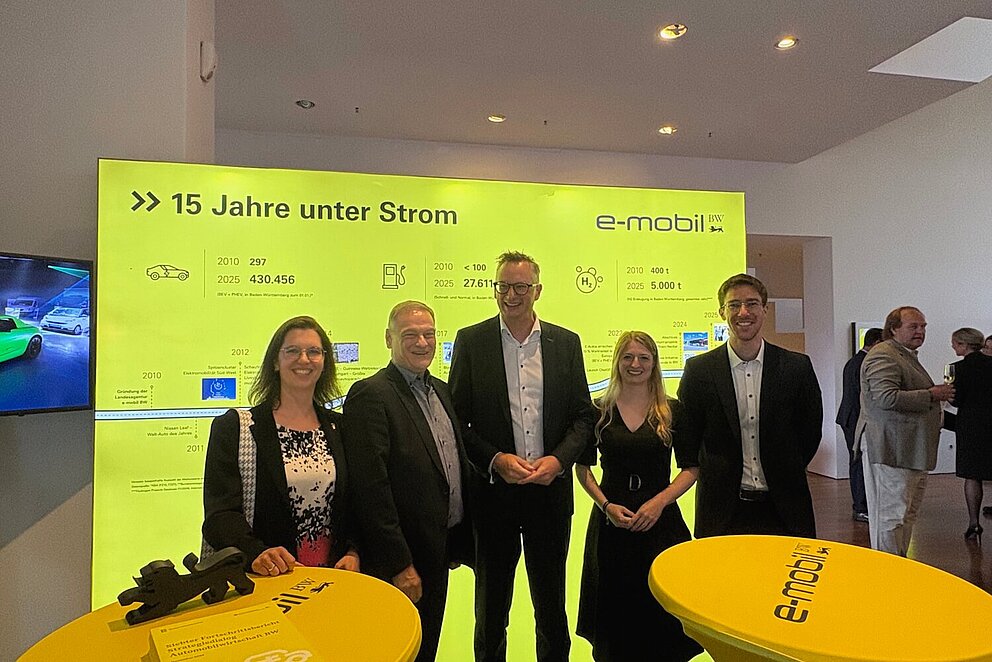 Eine Gruppe von fünf Personen steht lächelnd vor einem großen, leuchtend gelben Bildschirm mit der Aufschrift „15 Jahre unter Strom“ und dem Logo „e-mobil BW“. 