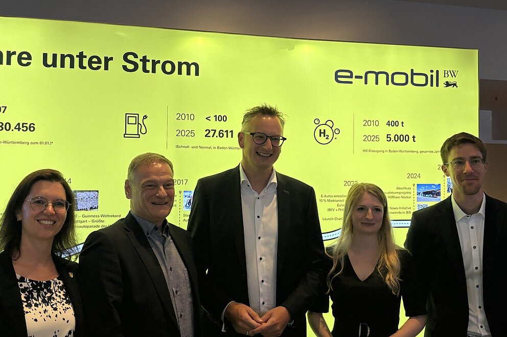 Eine Gruppe von fünf Personen steht lächelnd vor einem großen, leuchtend gelben Bildschirm mit der Aufschrift „15 Jahre unter Strom“ und dem Logo „e-mobil BW“. 