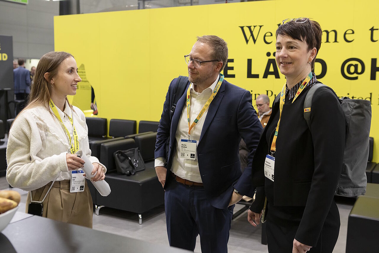 3 Personen im Gespräch vor einer Messewand.