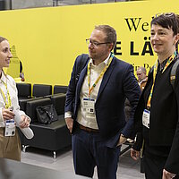 3 Personen im Gespräch vor einer Messewand.