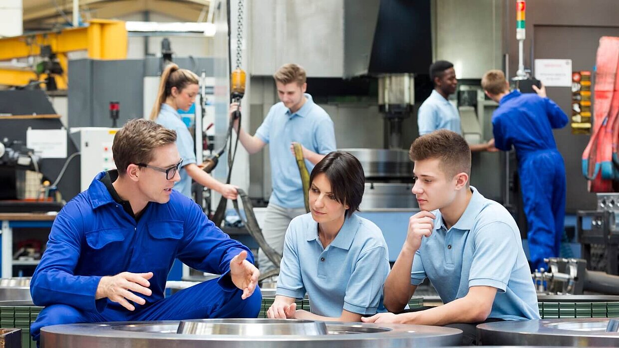 A man is teaching two younger people in a production hall.