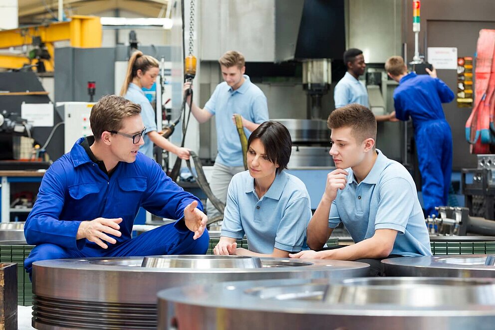 Ein Mann unterrichtet zwei jüngere Personen in einer Produktionshalle. 