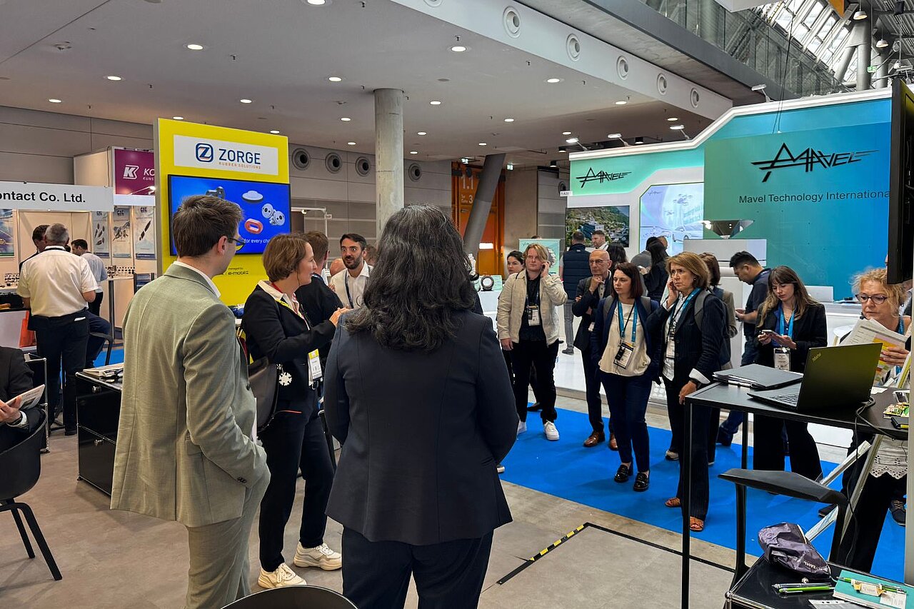 Eine Gruppe Personen informiert sich auf einem Messestand und hört über Kopfhörer einer Führung zu.