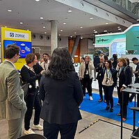 Eine Gruppe Personen informiert sich auf einem Messestand und hört über Kopfhörer einer Führung zu.