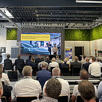 Blick auf die Bühne in der Zukunftswerkstatt 4.0 in Esslingen, Tim Siegel (e-mobil BW) moderiert durch die Veranstaltung, im Vordergrund sind die Teilnehmenden auf ihren Sitzplätzen zu sehen. 