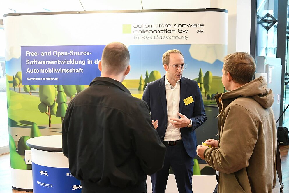 Ein Mitarbeiter der e-mobil BW steht vor einem Stand und berät zwei Interessierte auf einer Veranstaltung über das Thema „Free- and Open-Source-Software 