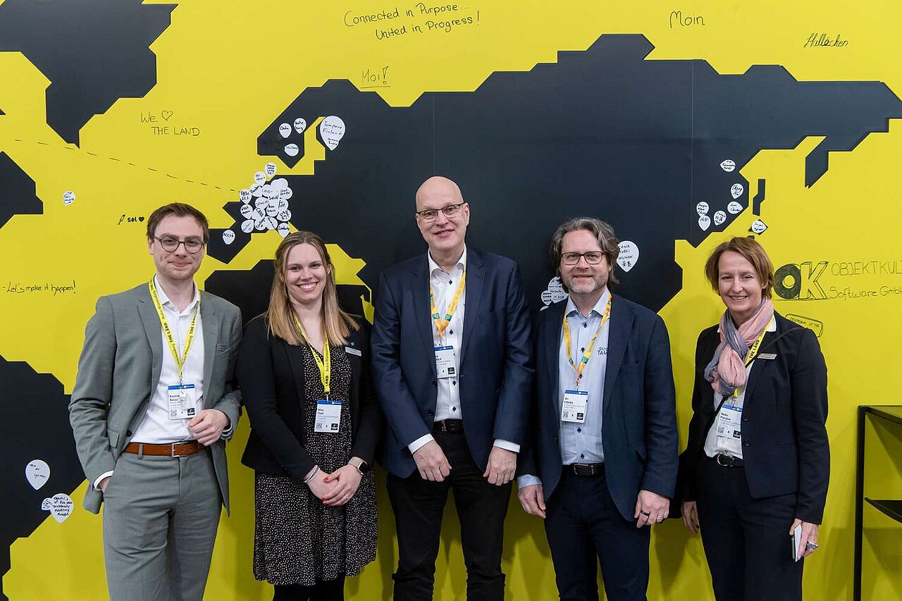 Fünf Menschen stehen vor einer gelben Wand mit schwarzem Aufdruck und lächeln.
