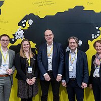 Fünf Menschen stehen vor einer gelben Wand mit schwarzem Aufdruck und lächeln.