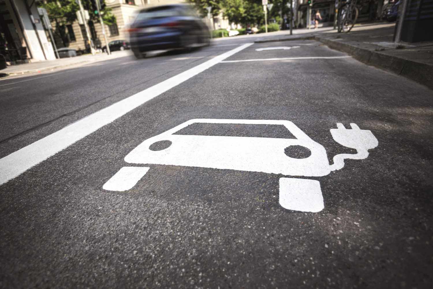 Auf der Straße ist im Vordergrund ein weißes Auto-Symbol mit Stecker zu sehen. Im Hintergrund fährt ein Auto vorbei.