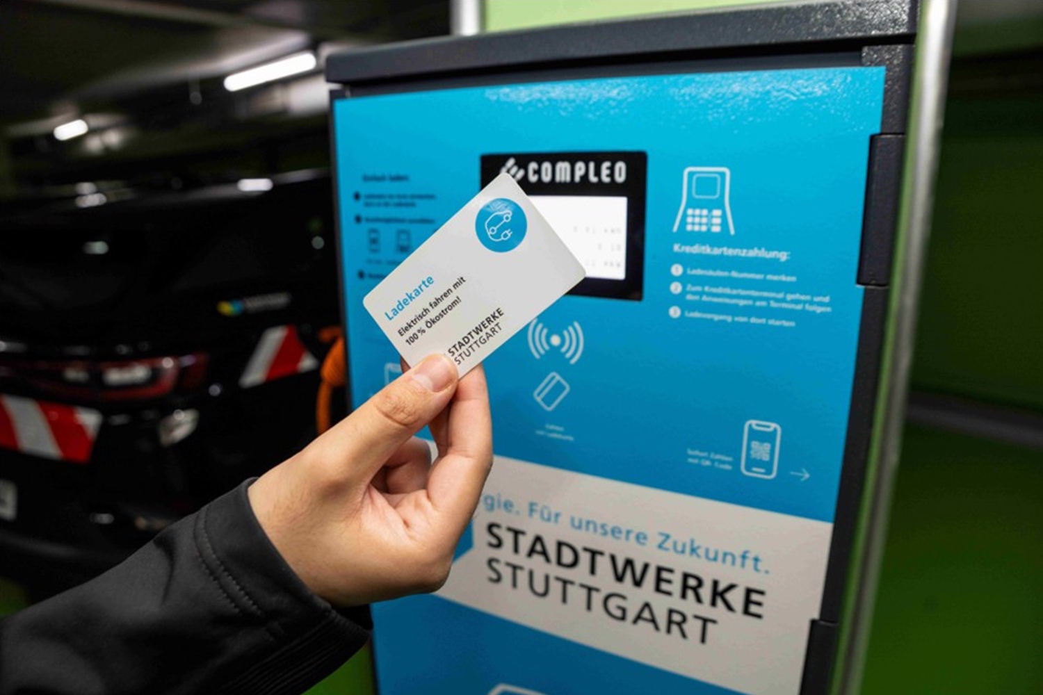 Eine Hand hält eine Ladekarte der Stadtwerke Stuttgart vor eine blaue Ladesäule.
