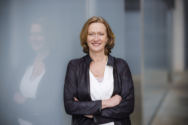 Kerstin Andreae lehnt mit verschränkten Armen an einer hellgrauen Glaswand