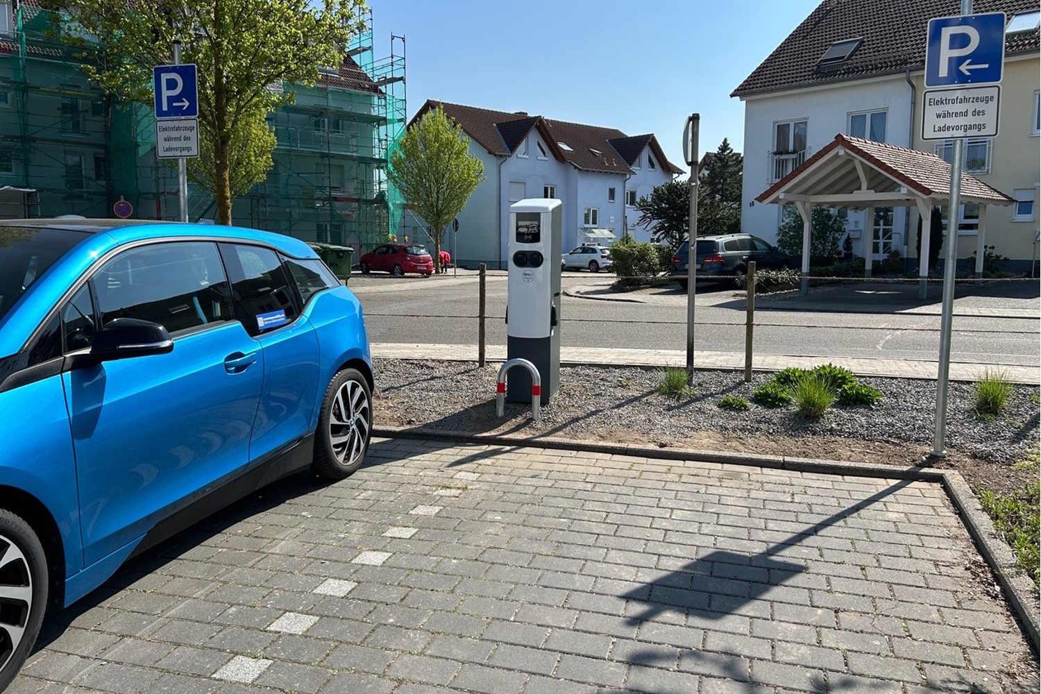 Ein blaues E-Auto steht auf einem Parkplatz neben einer Ladestation, dahinter sieht man ein Wohngebiet.
