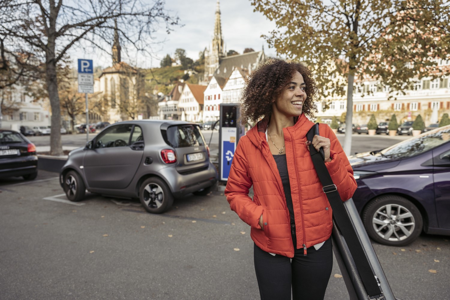 Lächelnde Frau im Vordergrund. Parkendes E-Auto an Ladestation im Hintergrund.