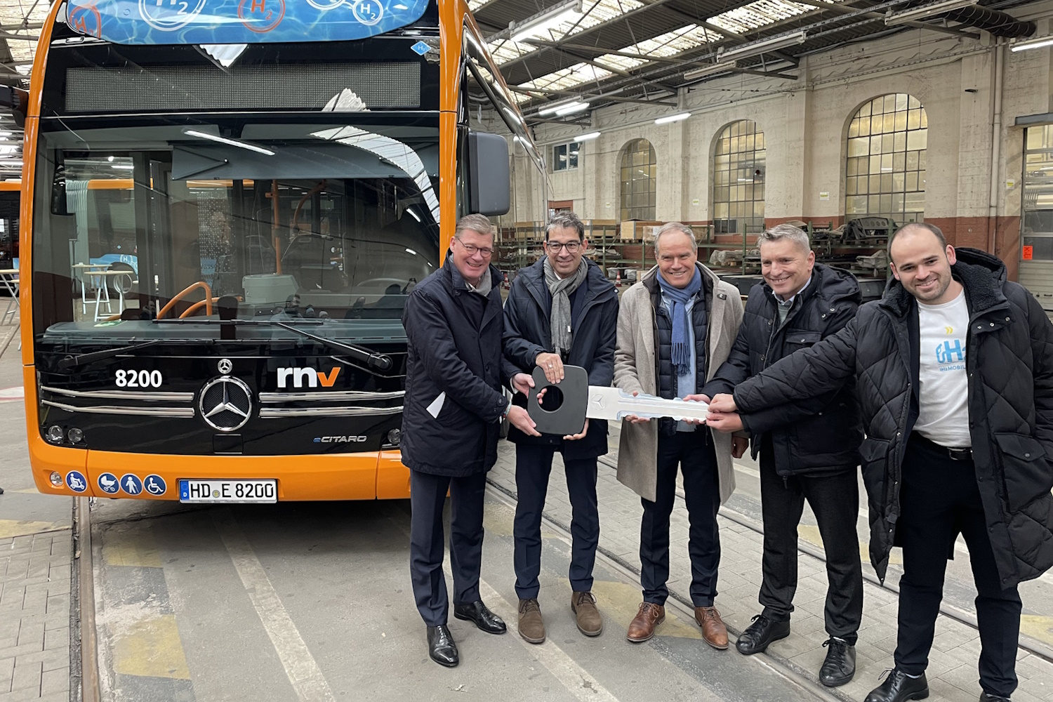 Eine gruppe Männer steht vor Wasserstoffbus in einer Halle und halten einen überdimensionierten Schlüssel in den Händen.