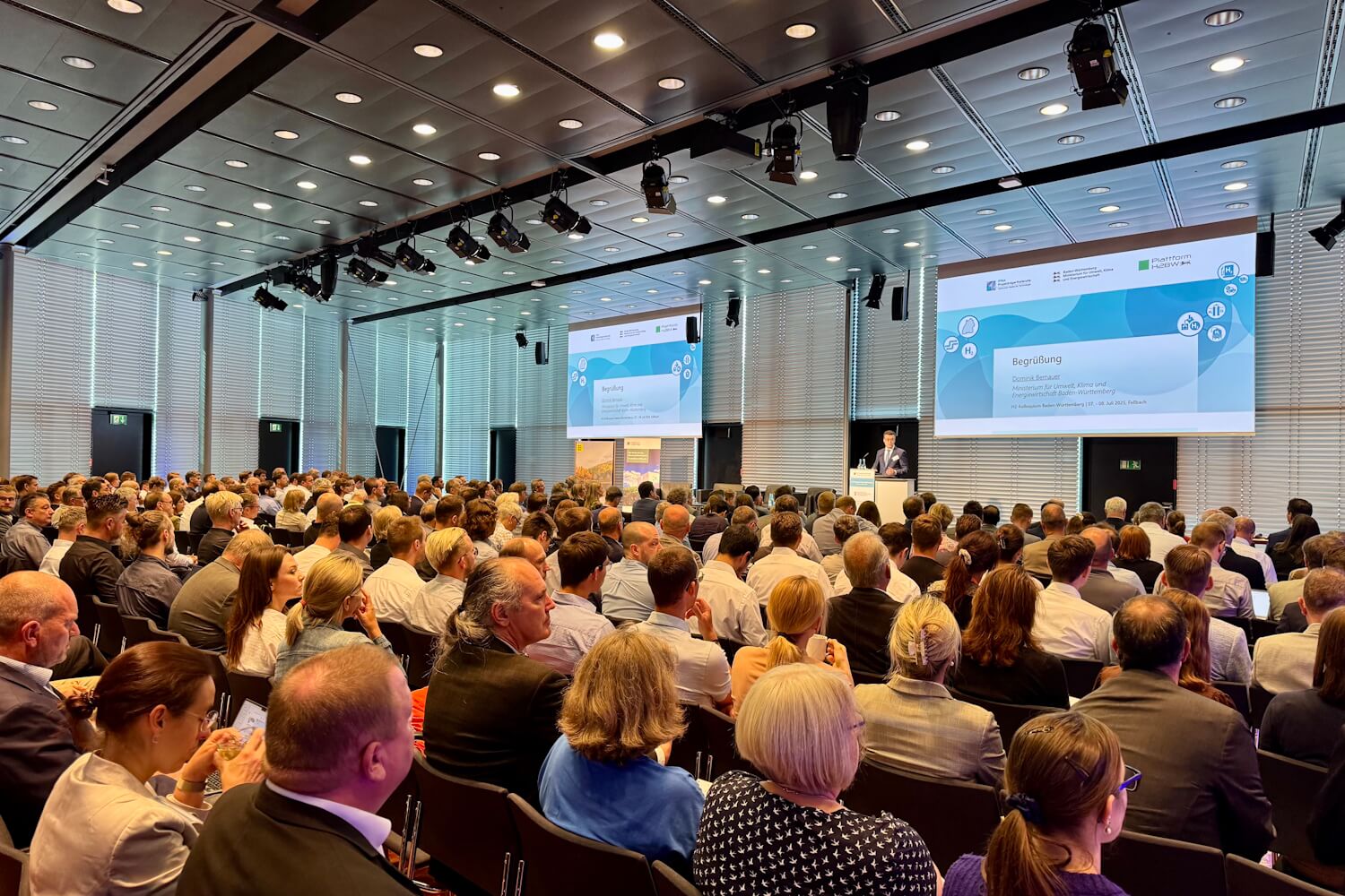 Großes Publikum bei einer Fachkonferenz in einem modernen Konferenzsaal, ein Redner steht am Podium, auf zwei Leinwänden ist die Begrüßungsfolie der Veranstaltung zu sehen.