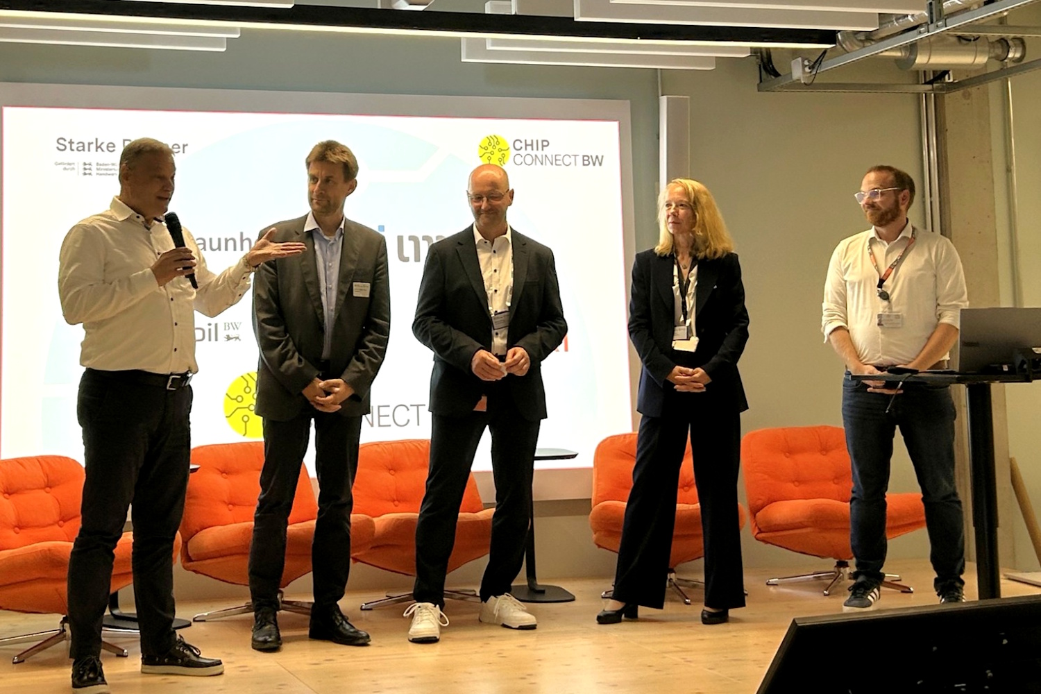 Fünf Menschen stehen auf einem Podium bei einer Veranstaltung. Ein Speaker hält das Mikrofon und spricht zum Publikum.