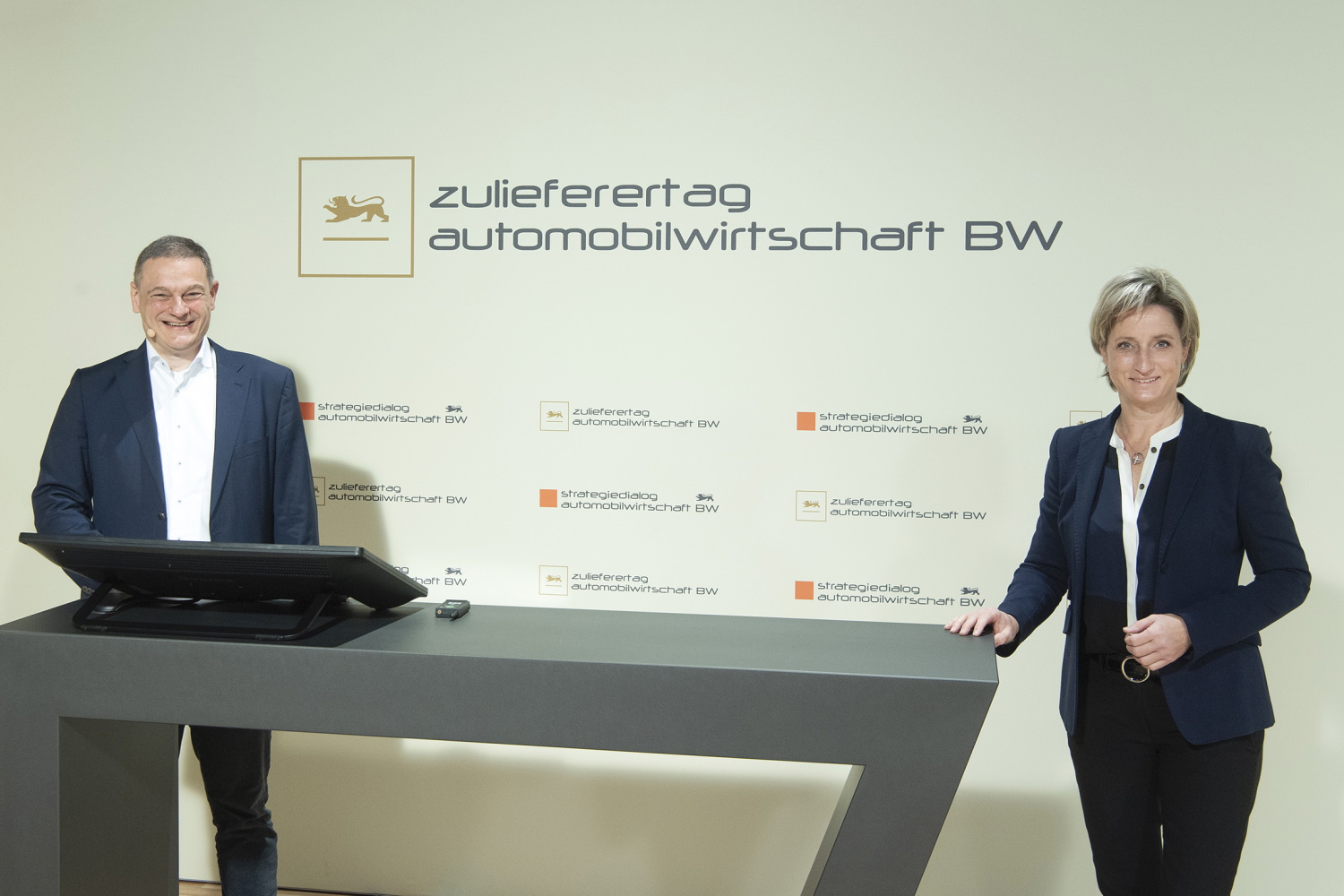 Zwei Personen stehen an einem Sprechpult und blicken lächelnd in die Kamera. Hinter ihnen ist das Logo des Zulieferertags Automobilwirtschaft BW zu sehen.