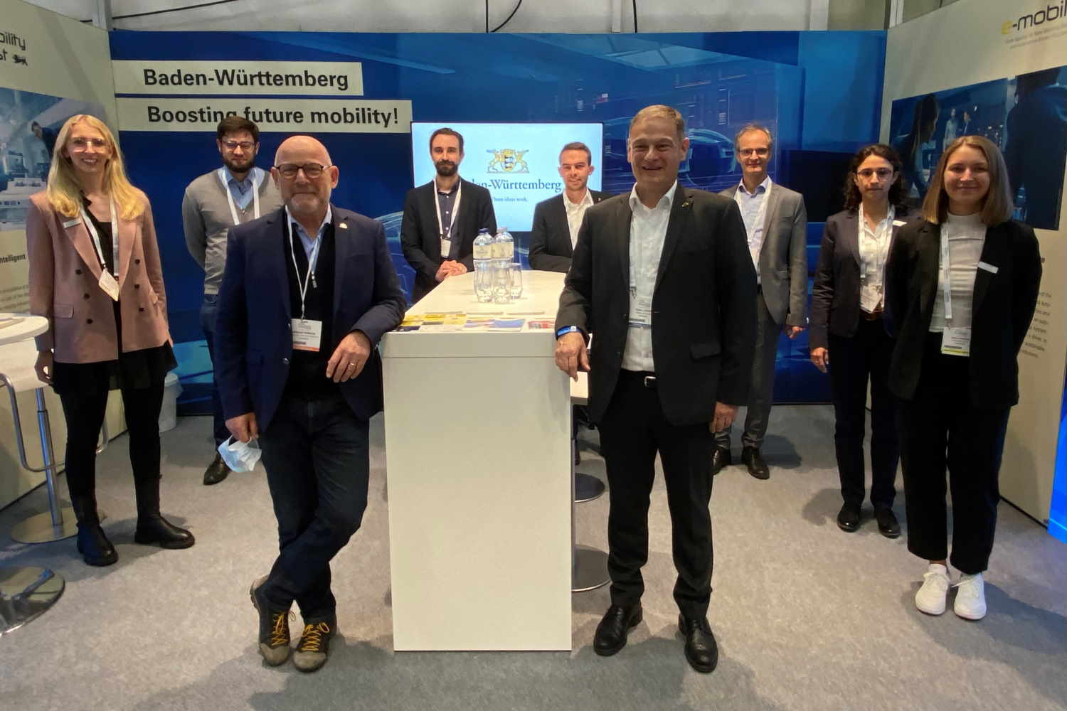 Gruppenbild am Messestand von Baden-Württemberg auf der ITS 2021. Zwei Personen in der Mitte vorne, angelehnt an einen Tisch. Im Hintergrund sieben weitere Kolleg:innen, die im Halbkreis angeordnet stehen und in die Kamera lächeln.