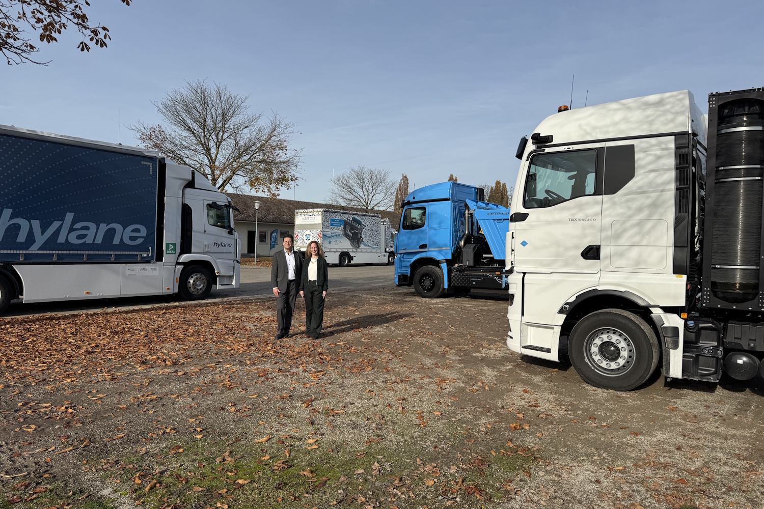 Vier Lkw stehen auf einer großen Fläche. In der Mitte stehen zwei Personen, die lächelnd in die Kamera blicken. 