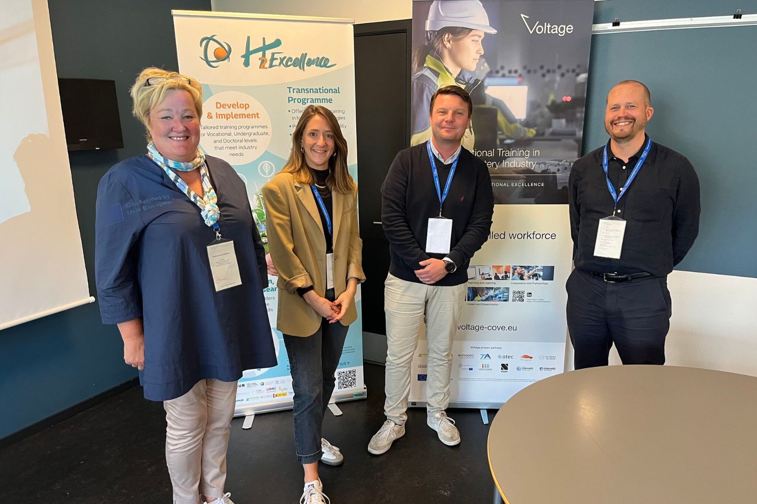 Vier Personen stehen in einem Raum während des Workshops im Rahmen des Forum on Vocational Excellence in Kolding, Dänemark. Links ein Banner des Projekts H2Excellence und rechts ein Banner des Projekts Voltage.